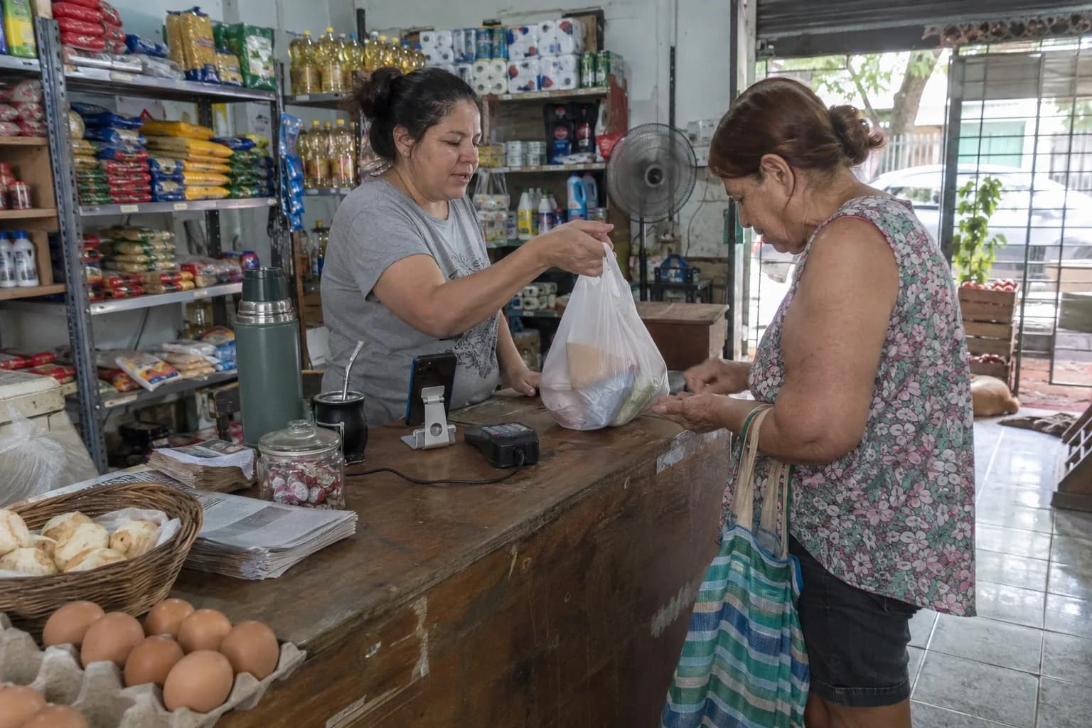 Almacenera entregando una bolsa de compras a una vecina en el mostrador de un almacén de barrio